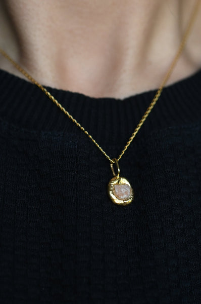 A close-up of a person wearing a delicate 24k vermeil gold chain necklace with a small, irregularly shaped gold pendant featuring a pale pinkish stone, set against a black knit sweater.