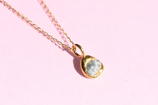 A gold chain necklace with a small, irregularly shaped gold plated silver pendant holding a pale blue-gray stone, displayed on a solid light pink background.