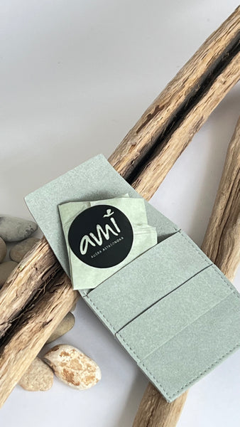 A sage green felt card holder wallet with a black circular Ami jewel sticker, styled on driftwood branches with small stones on a light grey background. Jewellery package 