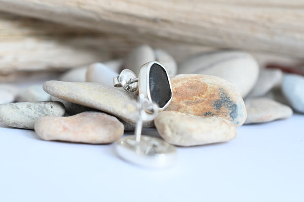 A silver stud earring with an irregular, black stone, displayed on smooth beach pebbles with a piece of driftwood in the background.