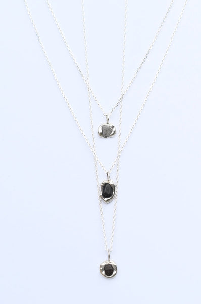 A layered necklace set featuring three delicate silver chains with small dark, organic-shaped pendants, displayed against a clean white background.