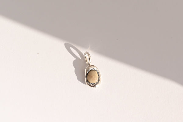 A small silver pendant with a textured, organic-shaped beige stone, photographed on a white surface with a strong diagonal shadow.