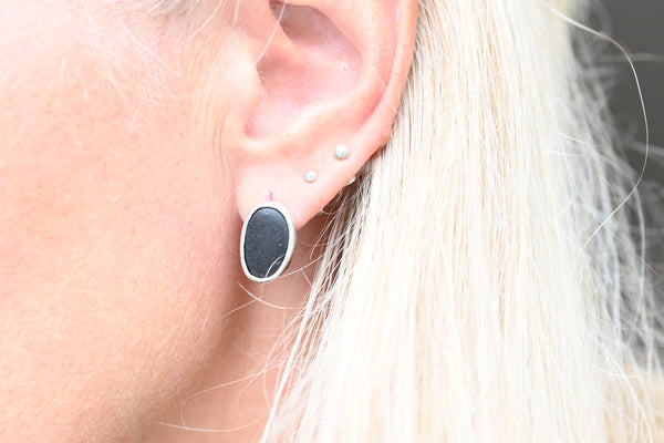 A close-up of a person's ear wearing a silver stud earring with an oval black stone, alongside two small pearl-like studs.