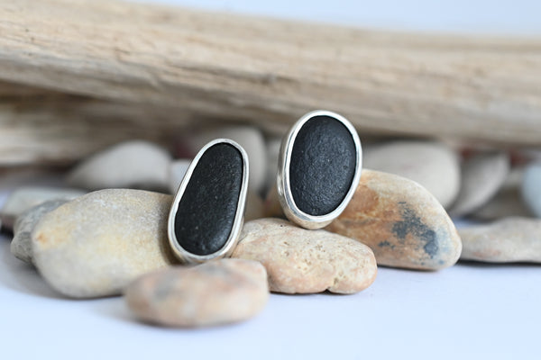 A pair of silver-toned oval stud earrings with dark, matte stone inlays, displayed on smooth river stones with a piece of driftwood in the background.