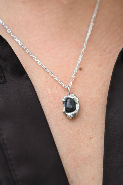A close-up of a person wearing a silver chain necklace with a dark black, irregularly shaped pendant, styled with a black satin top.
