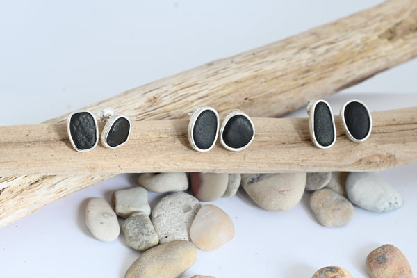 Three pairs of oval stud earrings with dark stone inlays in silver settings, displayed on driftwood with small stones on a white background.