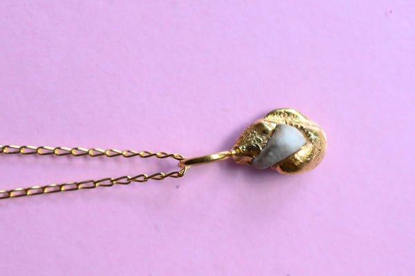 A gold chain necklace with a textured gold pendant featuring a white stone inlay, displayed on a solid pink background.