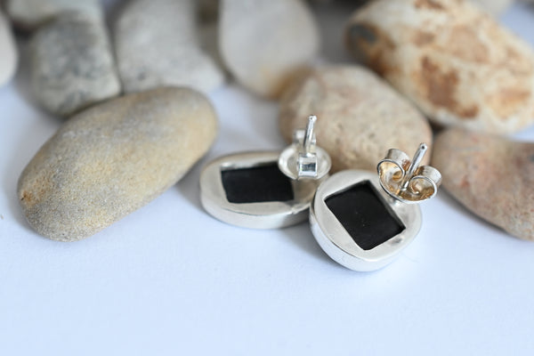 A pair of silver stud earrings with black seashore pebbles, displayed on a white surface with smooth stones in the background.