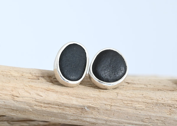 A pair of oval stud earrings with black seashore pebble in silver, displayed on a piece of driftwood with small stones below against a light blue background.