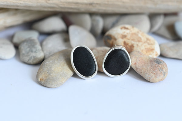 A pair of oval stud earrings with black stone inlays set in silver metal, displayed on smooth river stones against a white background.