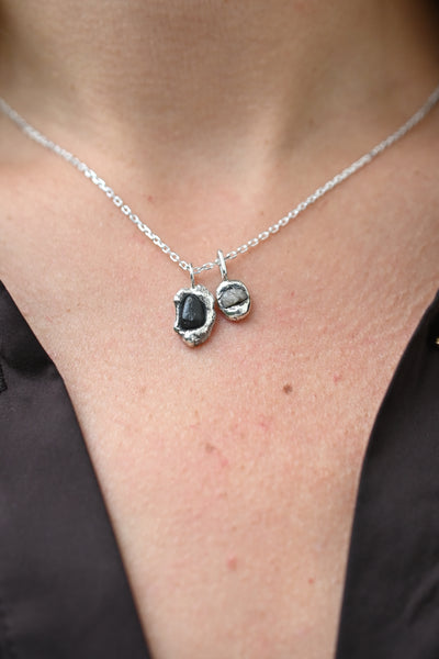 A close-up of a person wearing a silver necklace with two small pendants: one dark oval stone and one lighter gray stone, paired with a black satin top.