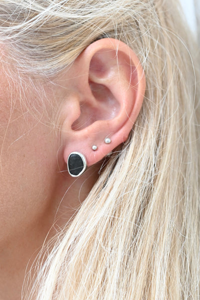 Close-up of a person's ear wearing a stack of earrings, including a large black oval stud with a silver bezel and two small pearl-like studs, with blonde hair framing the ear.