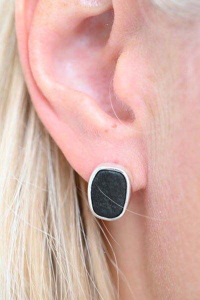 A close-up of a person's ear wearing a small stud earring with a dark rectangular stone set in a silver bezel, with blonde hair visible around it.