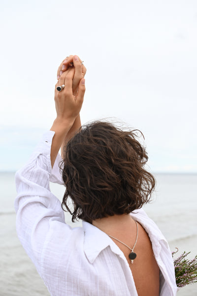 A woman seen from behind wearing a white linen shirt, with her back open to show a silver necklace with a dark round pendant with rose tourmaline. The background is a soft-focus beach and sky.