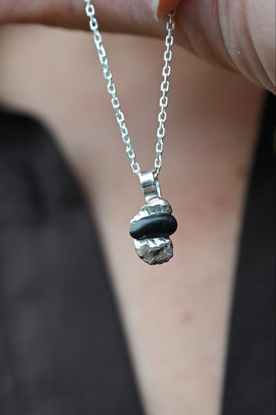 A close-up of a person holding a silver chain necklace with a unique pendant made of melted silver  and a dark black seashore stone  in the center.