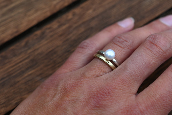 A close-up of a hand wearing a gold ring with a white pearl on a dark wooden surface.