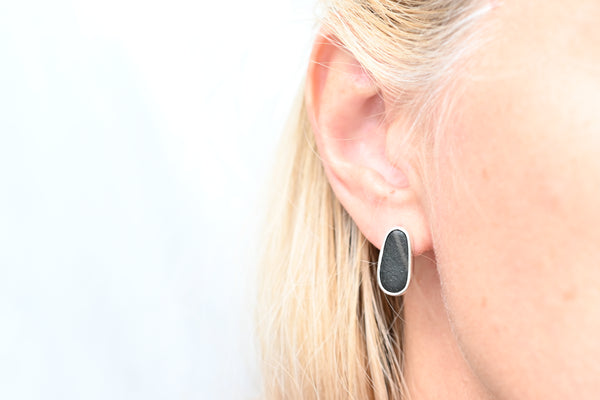 A close-up of a person's ear wearing a small teardrop-shaped black earring with a silver-bezel, against a bright white background.
