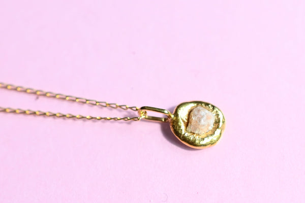 A 24k gold plated silver chain necklace with a small, irregularly shaped gold pendant featuring a pale, textured stone, displayed on a solid pink background.