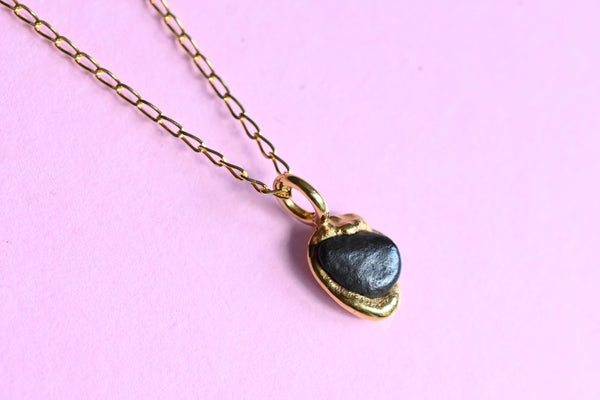 A gold chain necklace with a small heart-shaped pendant featuring a black Baltic sea stone, displayed on a solid light pink background.