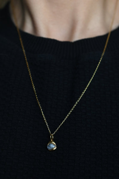 A delicate gold vermeil chain necklace with a small rounded blue-gray stone pendant, worn by a person in a black knit top.