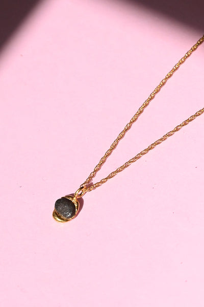 A delicate gold chain necklace with a small bezel-set dark brown stone pendant, displayed on a solid pink background with a soft shadow.