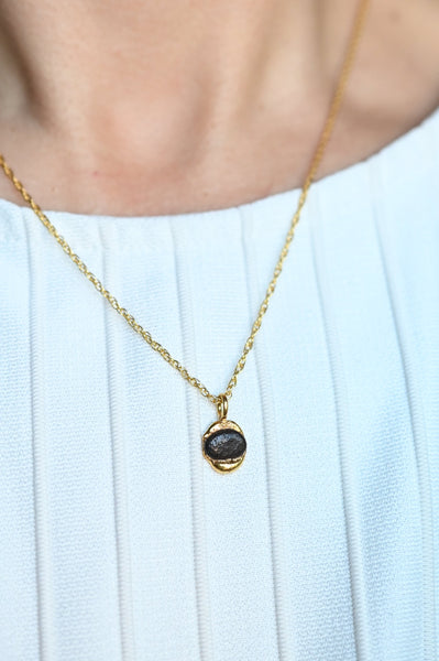 A close-up of a person wearing a delicate gold chain necklace with a small oval dark seashore stone pendant, paired with a white ribbed top.