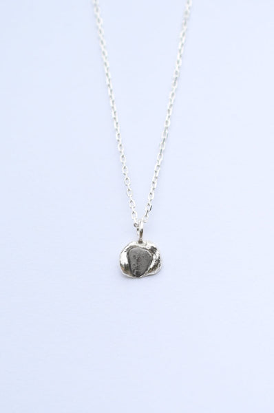 A silver chain necklace with a small, textured, organic-shaped pendant, displayed against a light blue background.