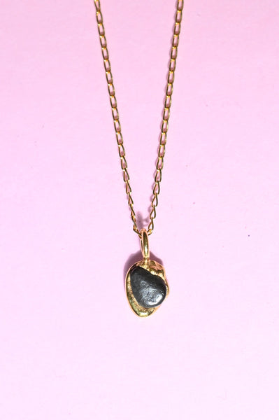 A gold chain necklace with a small heart-shaped pendant featuring a black natural stone, displayed on a solid pink background.