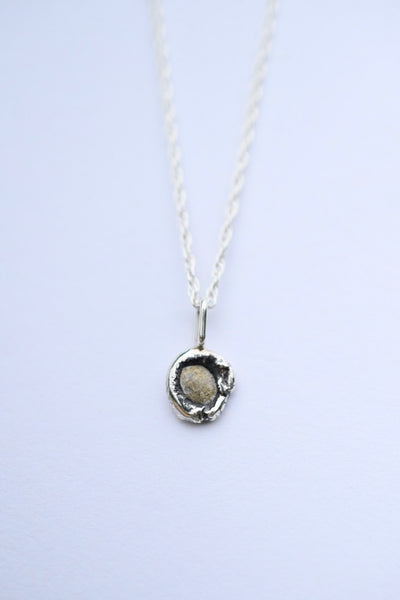 A silver necklace with a textured, organic-shaped pendant containing a small beige stone, suspended on a delicate silver chain against a light lavender background.