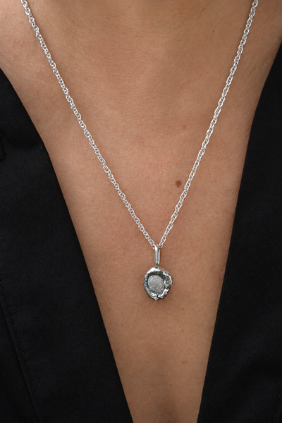 A close-up of a person's neck and chest wearing a delicate silver chain necklace with a small round bezel-set gray stone pendant, paired with an oxidised sterling silver.