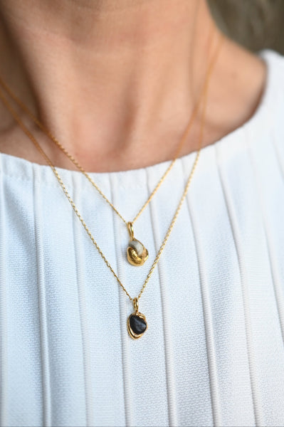 A close-up of a person wearing two delicate gold necklaces over a white pleated top. The necklaces feature small organic-shaped pendants, one in white and one with a dark, polished stone in a gold setting.
