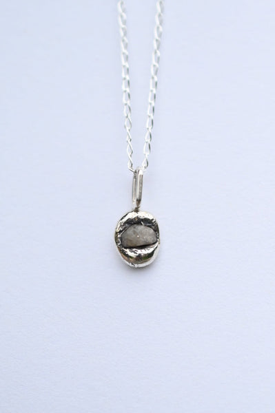 A silver chain necklace with a small, rough, oval-shaped gray stone pendant, displayed against a light gray background.