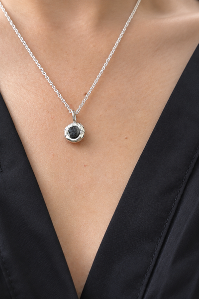 A close-up of a person wearing a delicate silver chain necklace with a textured silver pendant holding a dark, round stone, styled with a black V-neck top.