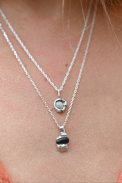 A close-up of a person wearing two layered silver necklaces. The shorter chain features a small round beige stone pendant, while the longer chain holds a larger silver capsule-shaped pendant with a dark beach stone.