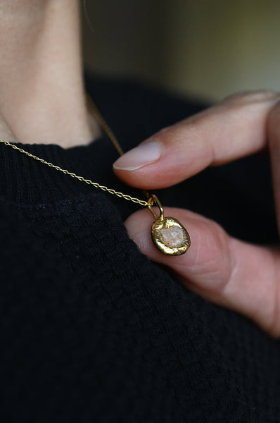A close-up of a person wearing a delicate gold chain necklace with a small, textured gold oval pendant, held gently by a hand against a black knit top.