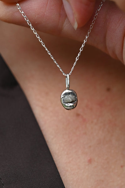 A close-up of a person holding a silver chain necklace with a small, textured oval pendant with natural grey stone against their neck and chest.