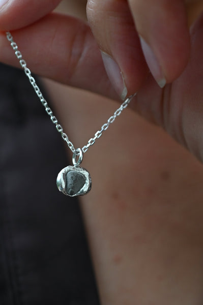 A close-up of a hand holding a delicate silver chain necklace with a small, textured grey stone pendant.